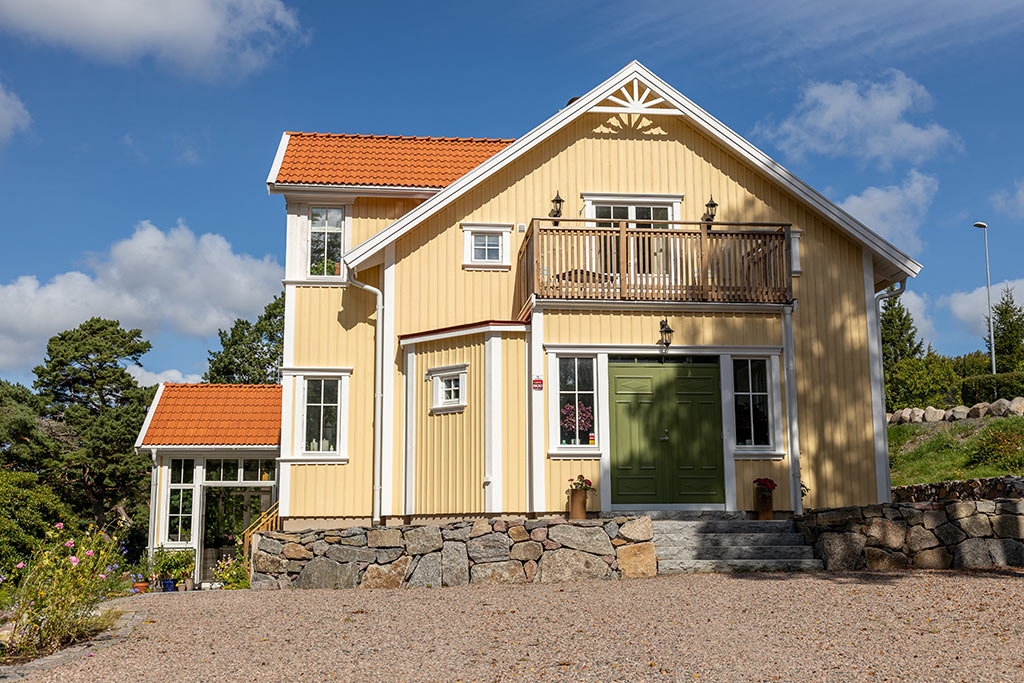 Det gula huset med vita knutar pryds av en ytterdörr i en grön kulör. På taket ligger tvåkupigt lertegel.